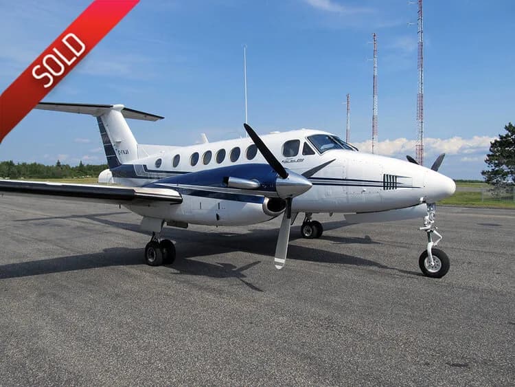 A white and blue Beechcraft King Air 200 series aircraft parked on an asphalt tarmac under a clear sky.