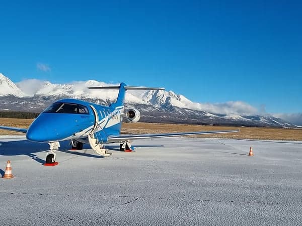 A blue and white Pilatus PC-24 on a snowy airfield with mountains in the background.