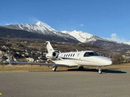 A white private jet with registration F-GNBY parked on a runway with mountains in the background.