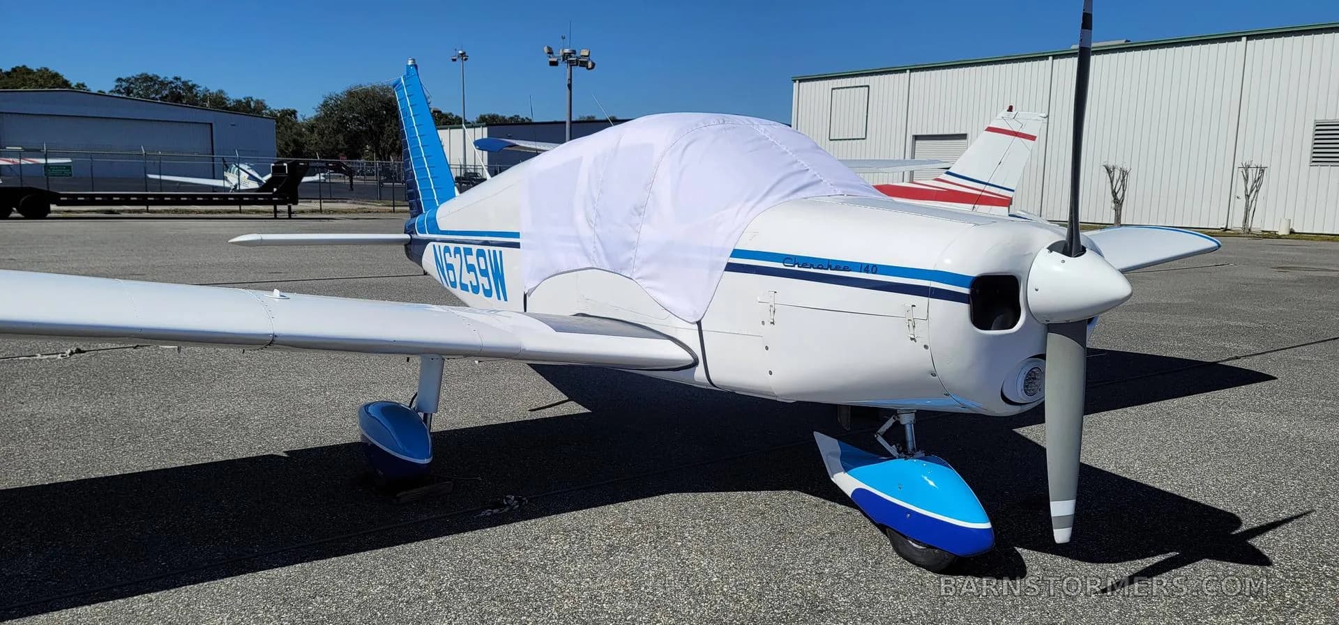 A white and blue light aircraft with a white cockpit cover parked on an airfield under a clear sky.