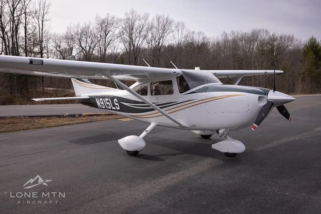 A white and dark green Cessna Skylane N915LS with gold stripes, parked on an asphalt ramp with trees in the background.
