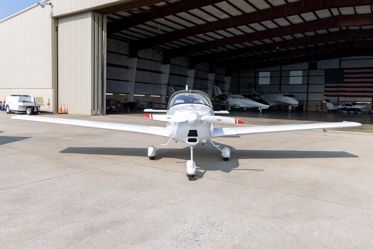 A white Tecnam Astore light sport aircraft with blue and grey stripes viewed from the front, parked on a tarmac.