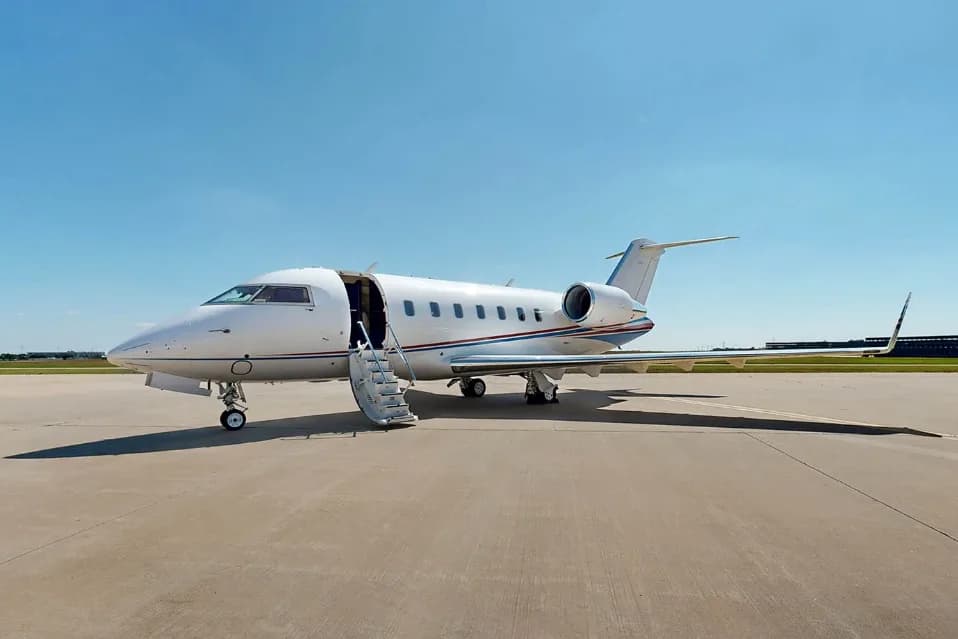 A white Bombardier Challenger business jet with blue and red accent stripes is parked on a tarmac with its passenger door open and stairs extended.