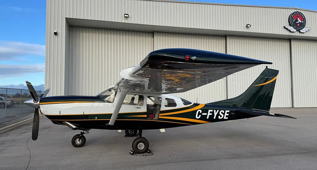 A side view of a dark green and white Cessna 182 with yellow stripes, registration C-FYSE, parked on an airfield.