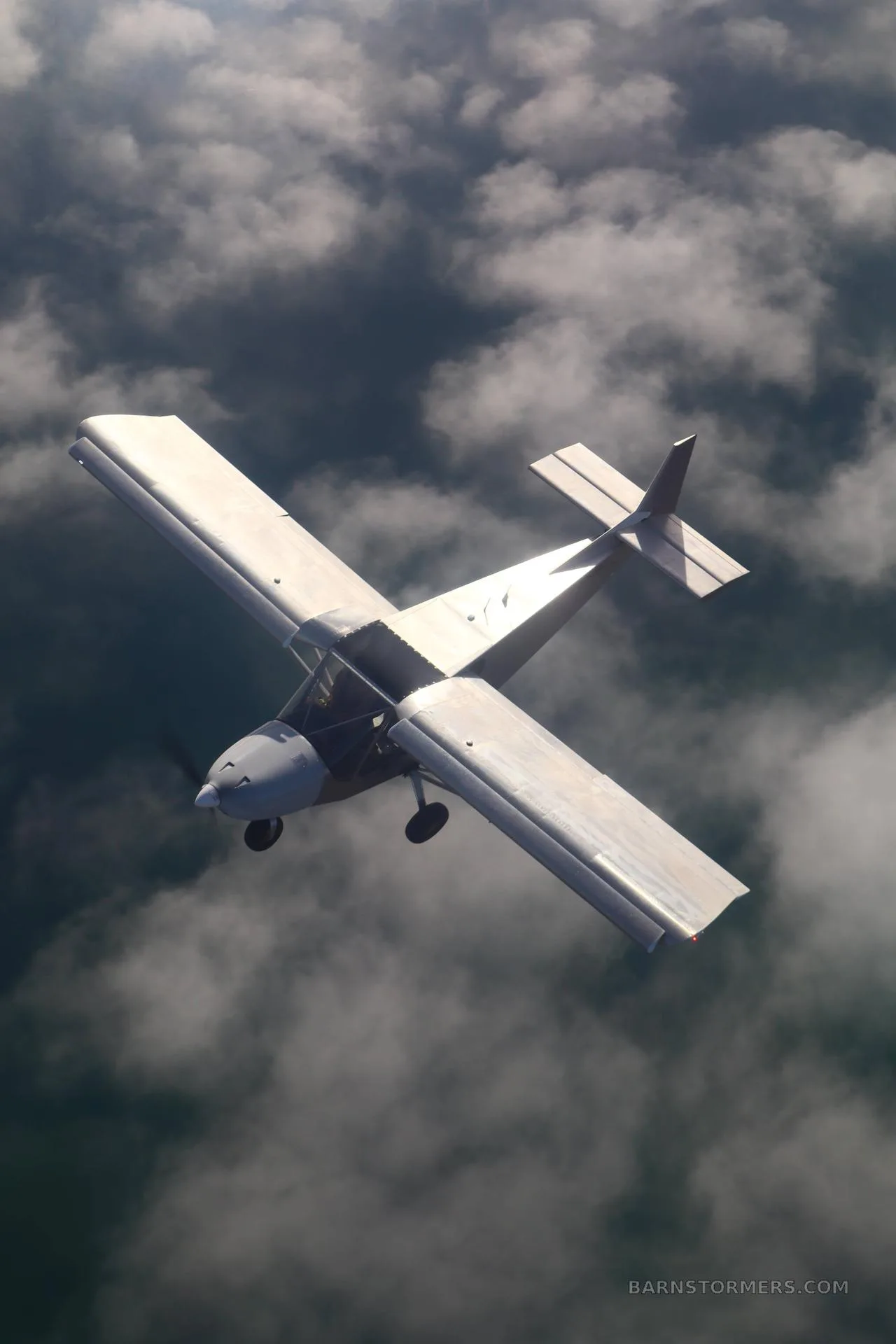 An aerial view of a light, high-wing aircraft with a silver finish flying above a layer of dark clouds.