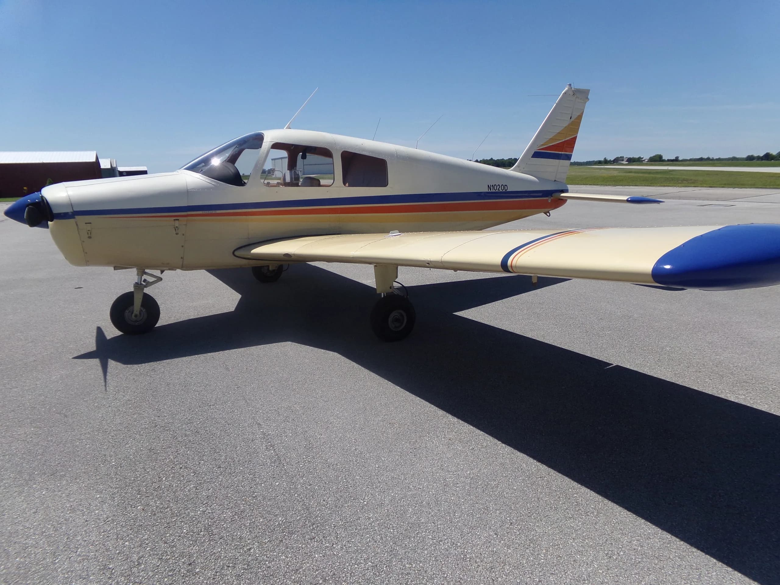 A white and beige Piper aircraft with blue, orange, and yellow stripes on the fuselage and tail, parked on an asphalt tarmac under a clear blue sky.