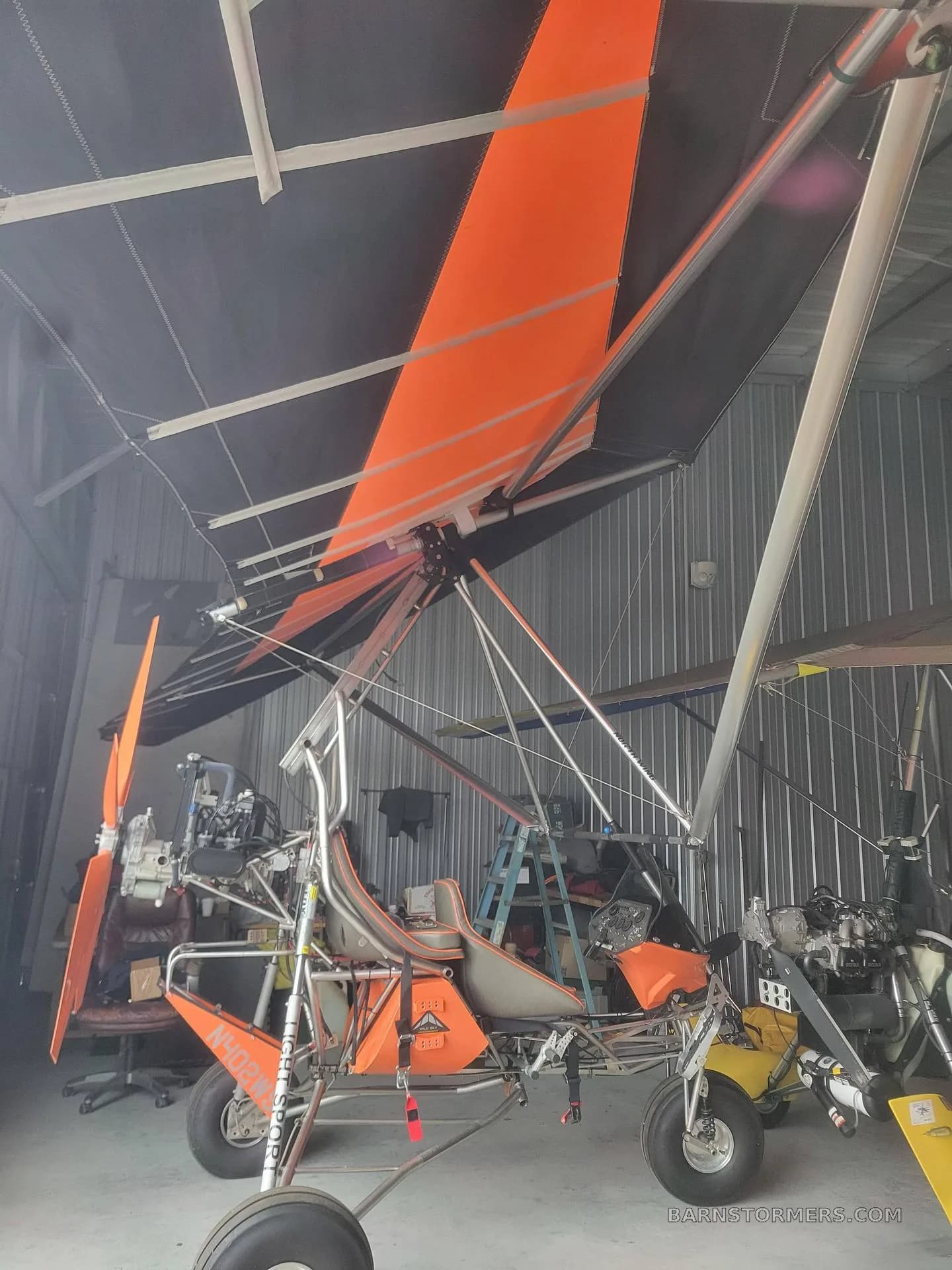 An orange and black weight-shift control ultralight aircraft parked inside a hangar, surrounded by other aircraft parts and equipment.