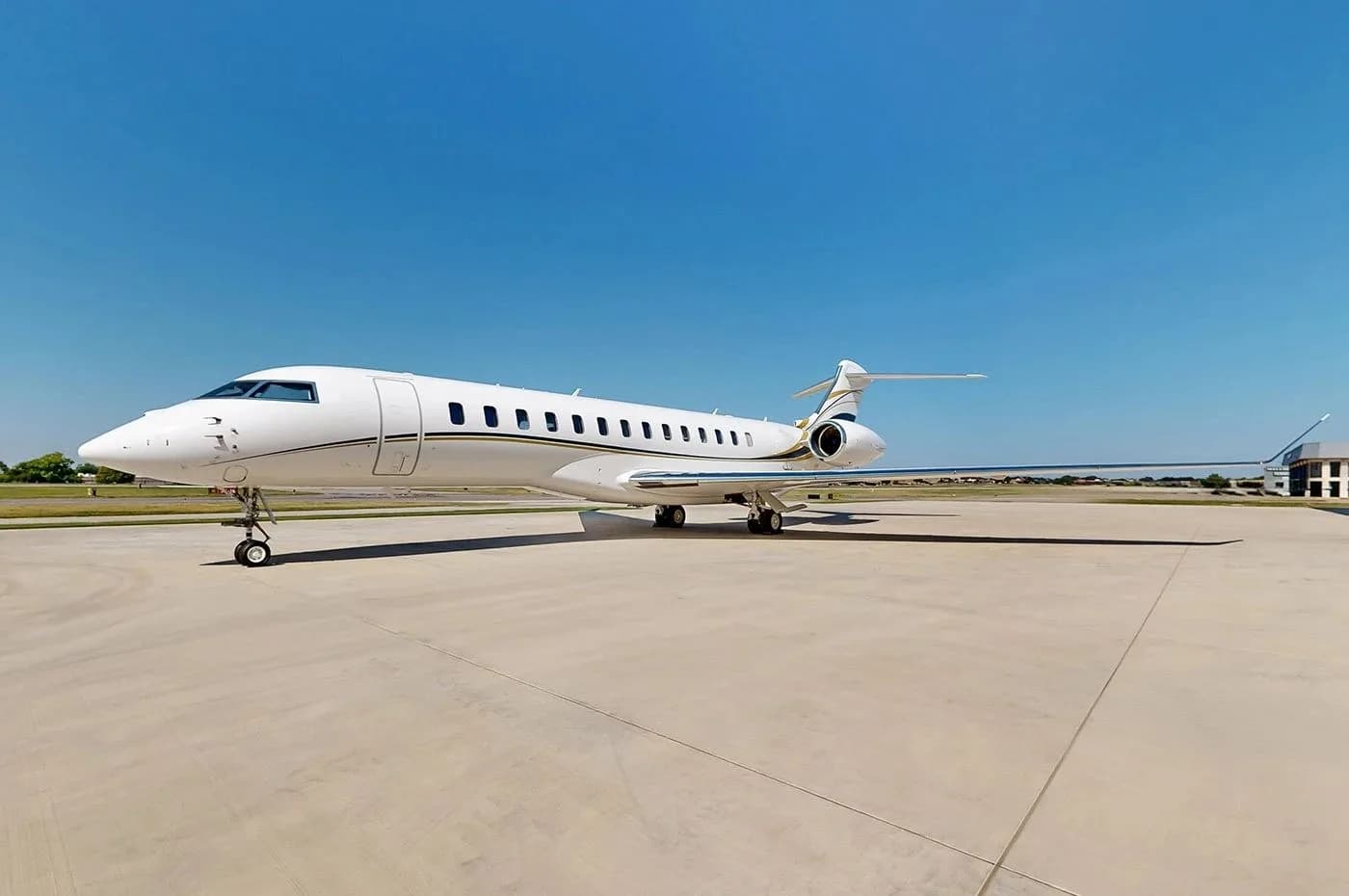 A white Bombardier Global business jet with gold and black stripes parked on a tarmac under a clear blue sky.