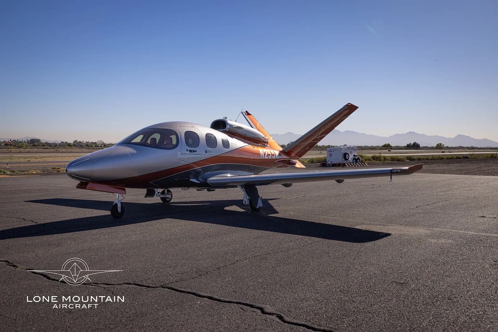 A three-quarter front view of a silver and orange Cirrus Vision Jet (SF50) parked on an asphalt tarmac with mountains in the distance.