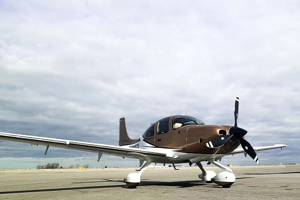 A Cirrus SR22 aircraft with a brown and white paint scheme parked on an airfield under a cloudy sky.