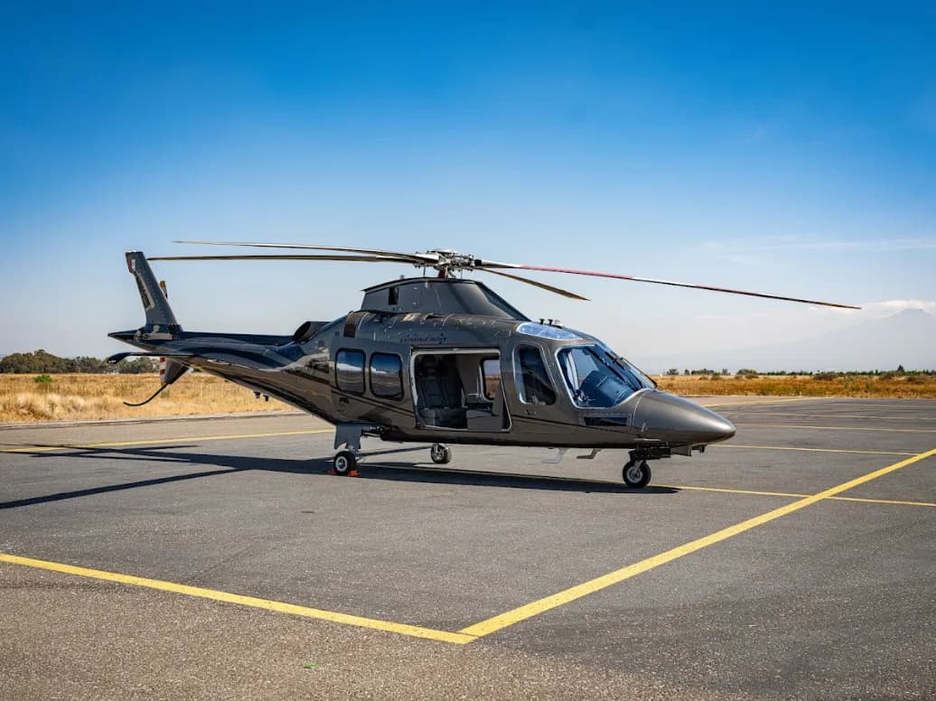 A grey AgustaWestland AW109 GrandNew helicopter with an open passenger door is parked on an airfield under a clear blue sky.