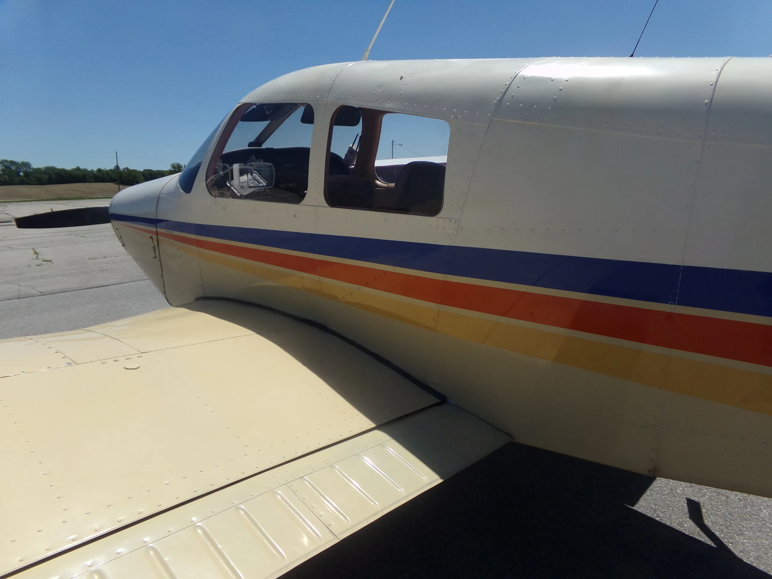 A close-up view of the cockpit windows and front fuselage of a Piper aircraft, showing the blue and orange stripe.