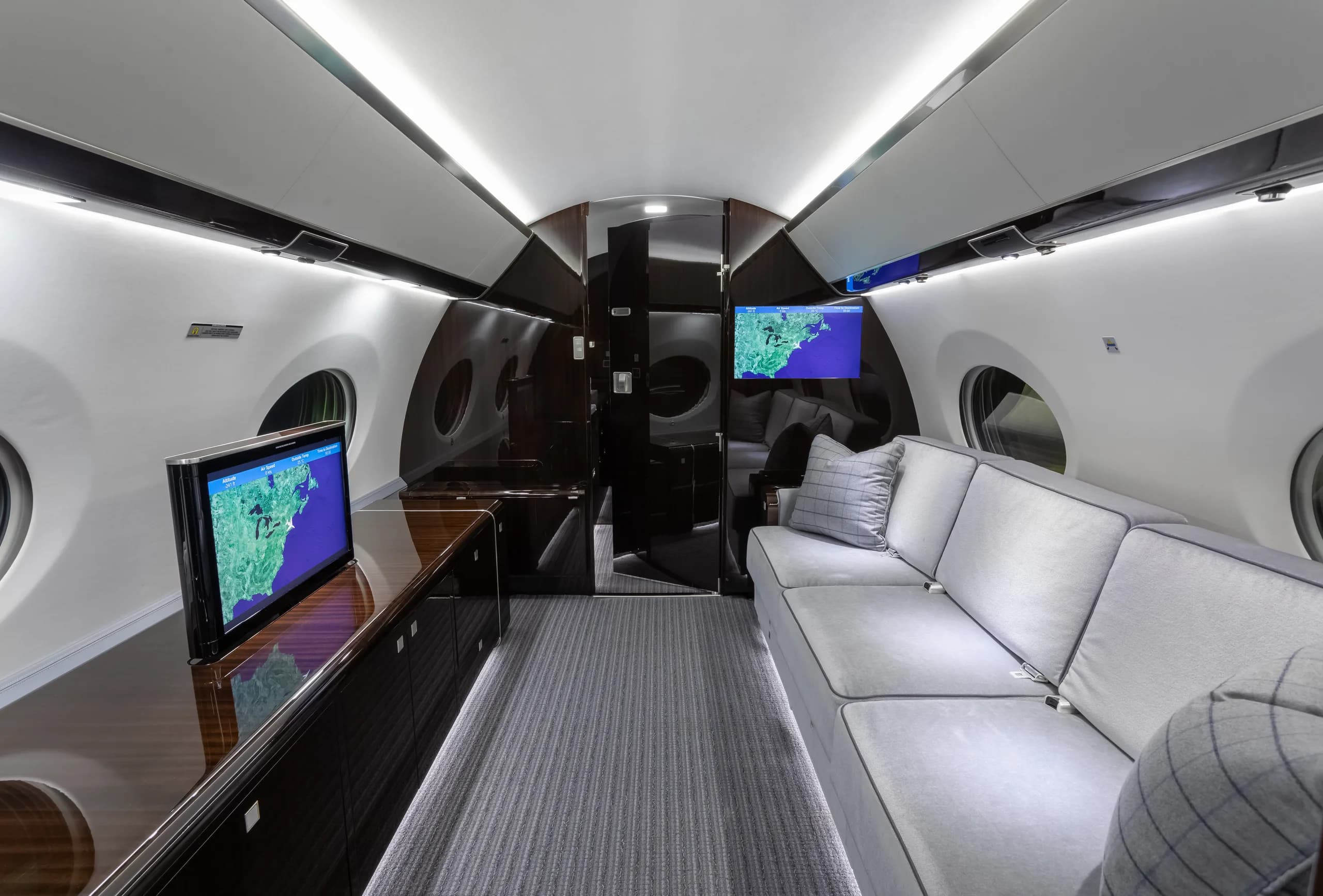 The aft section of the private jet cabin, featuring a long grey sofa, dark wood cabinetry, and a large screen displaying a map.