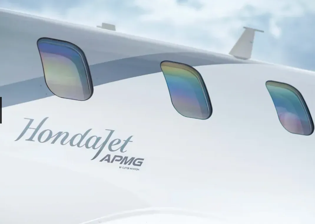 A close-up of the HondaJet fuselage, showing three cabin windows with rainbow reflections and the "HondaJet APMG" branding.