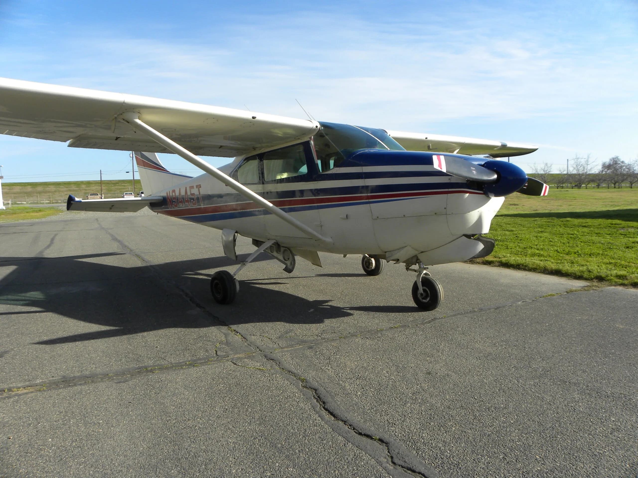 A white Cessna 182 with blue and red stripes parked on a tarmac with grass in the background.