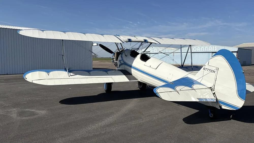 A white and blue biplane, N777CH, parked on an airfield with hangars in the background.
