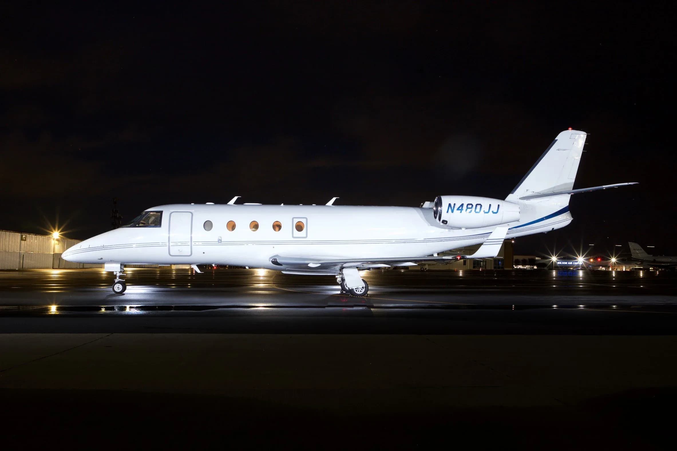 A white business jet with blue tail stripes is parked on a wet tarmac at night, illuminated by ground lights.