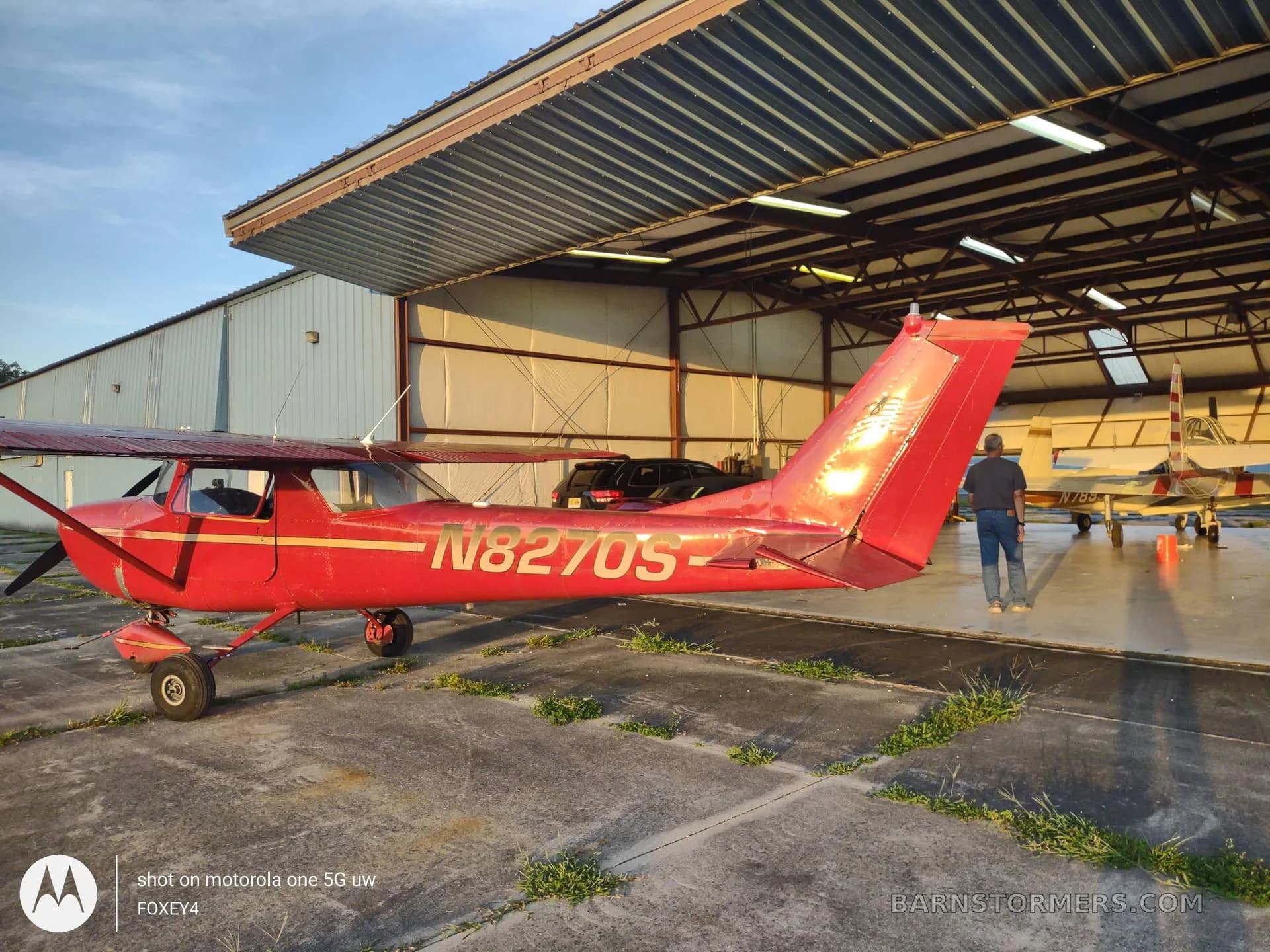 A red Cessna 150 or similar light aircraft with registration N8270S is parked on a concrete apron, partially in front of an open hangar.