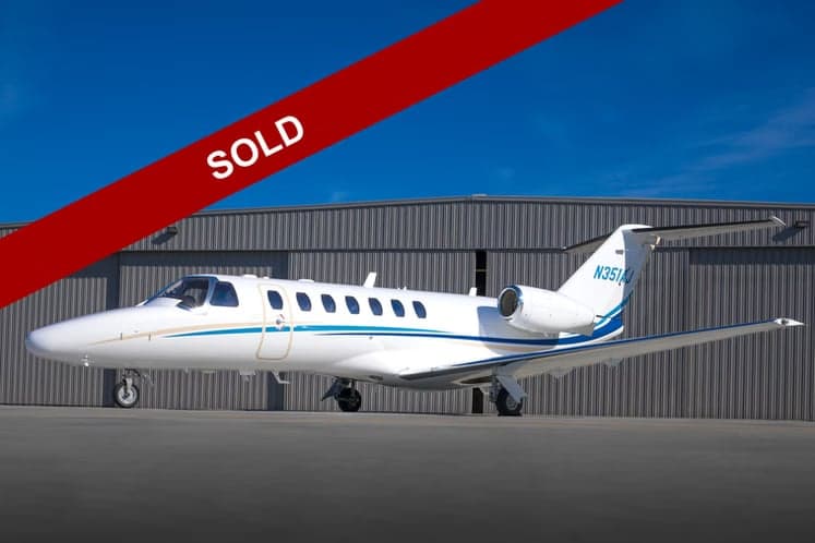 A white Cessna Citation CJ3 aircraft with blue and gold accents is parked in front of a hangar, with a red 'SOLD' banner across the image.