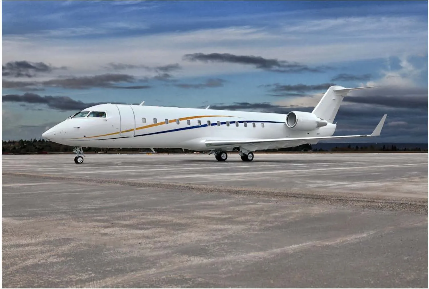 A white business jet with blue and gold stripes on an airfield under a cloudy sky.