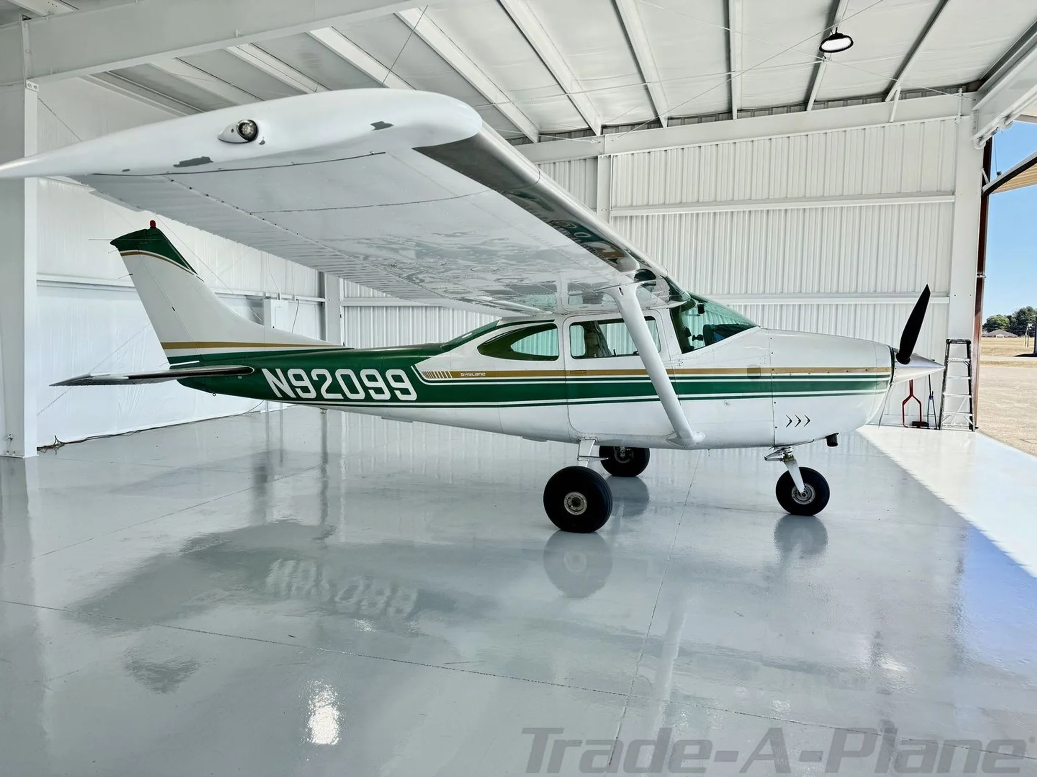 A white and green Cessna Skylane N92099 parked inside a hangar with the door open, revealing a sunny sky outside.