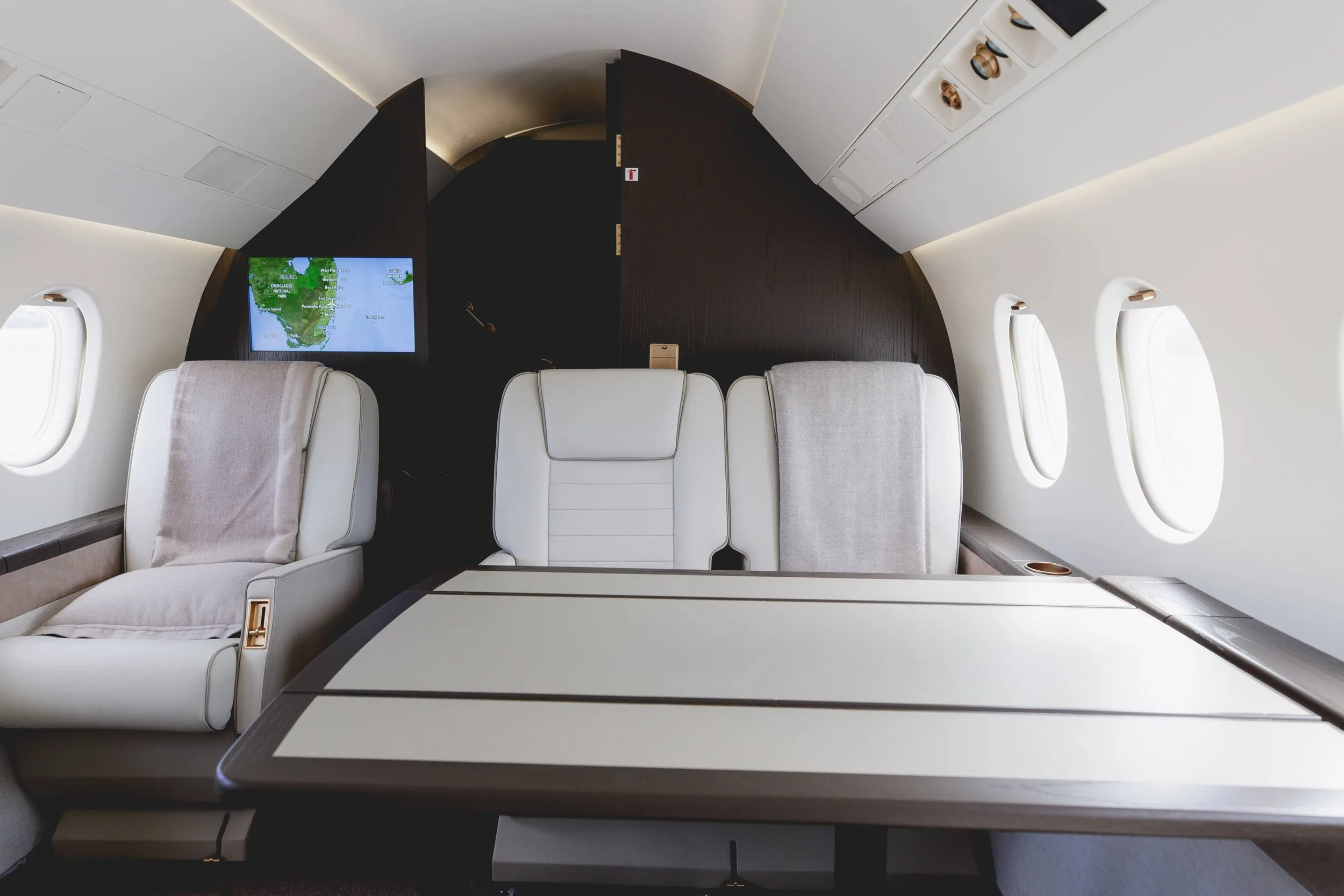 A view of the aircraft cabin showing a conference table and two facing white leather seats with a map displayed on a screen.