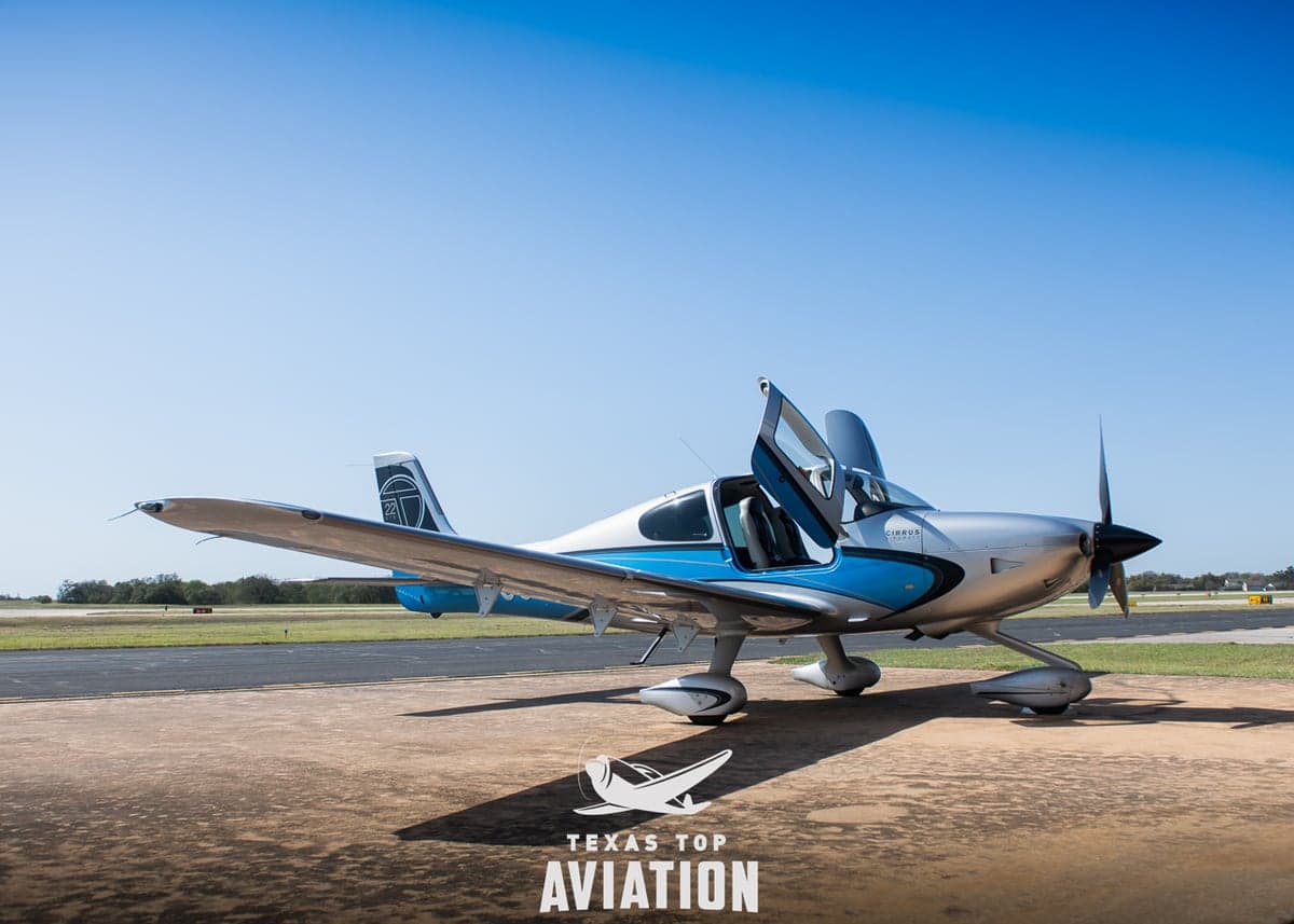 A Cirrus aircraft is parked on the tarmac with its doors open, featuring a silver and blue paint scheme.