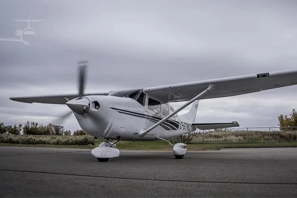 A white single-engine propeller aircraft, registration C-GTMQ, with a spinning propeller, parked on an asphalt runway under an overcast sky.