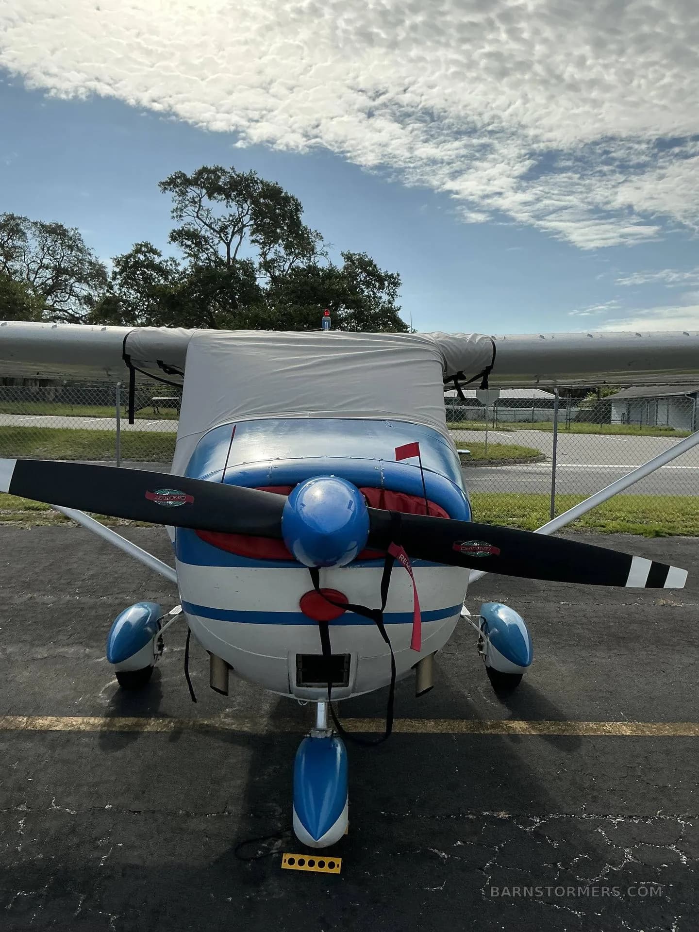 A white and blue Cessna 172 aircraft with a grey cockpit cover and red propeller flags is viewed from the front.