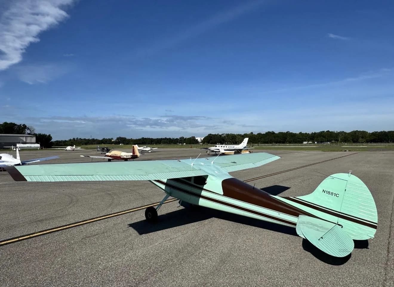 An aerial-like view from behind a brown and light green Cessna 170B (N1881C) on an airport runway with other aircraft in the background.
