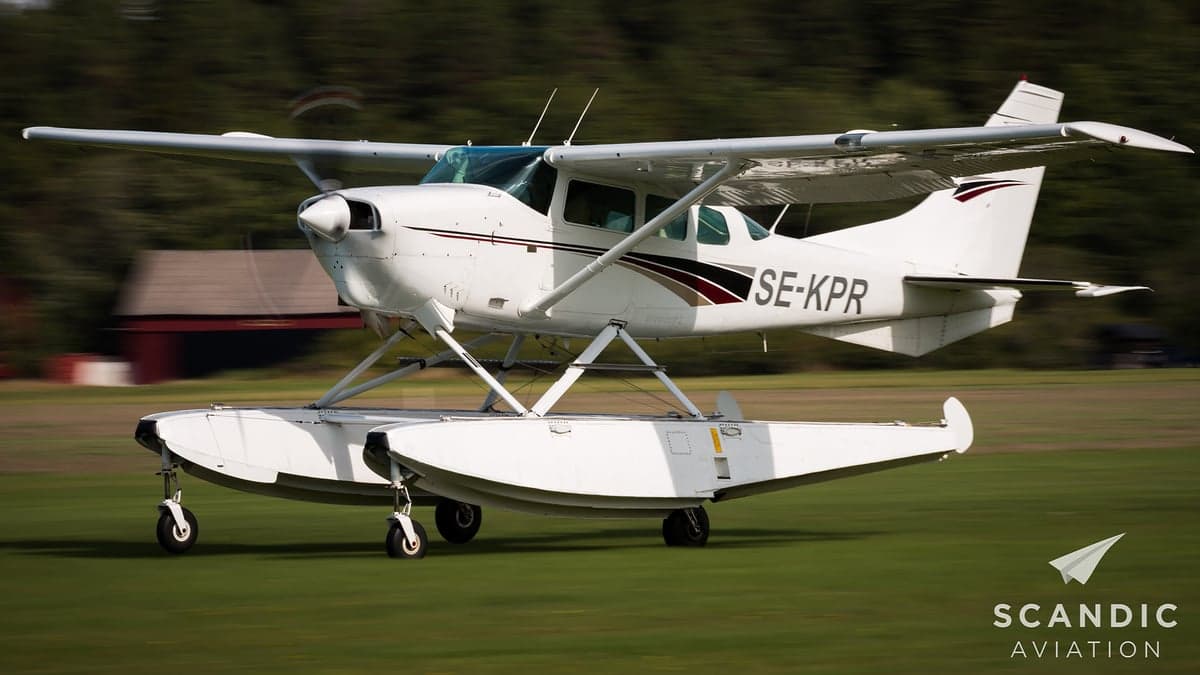 White and maroon floatplane taxiing on a grassy field.