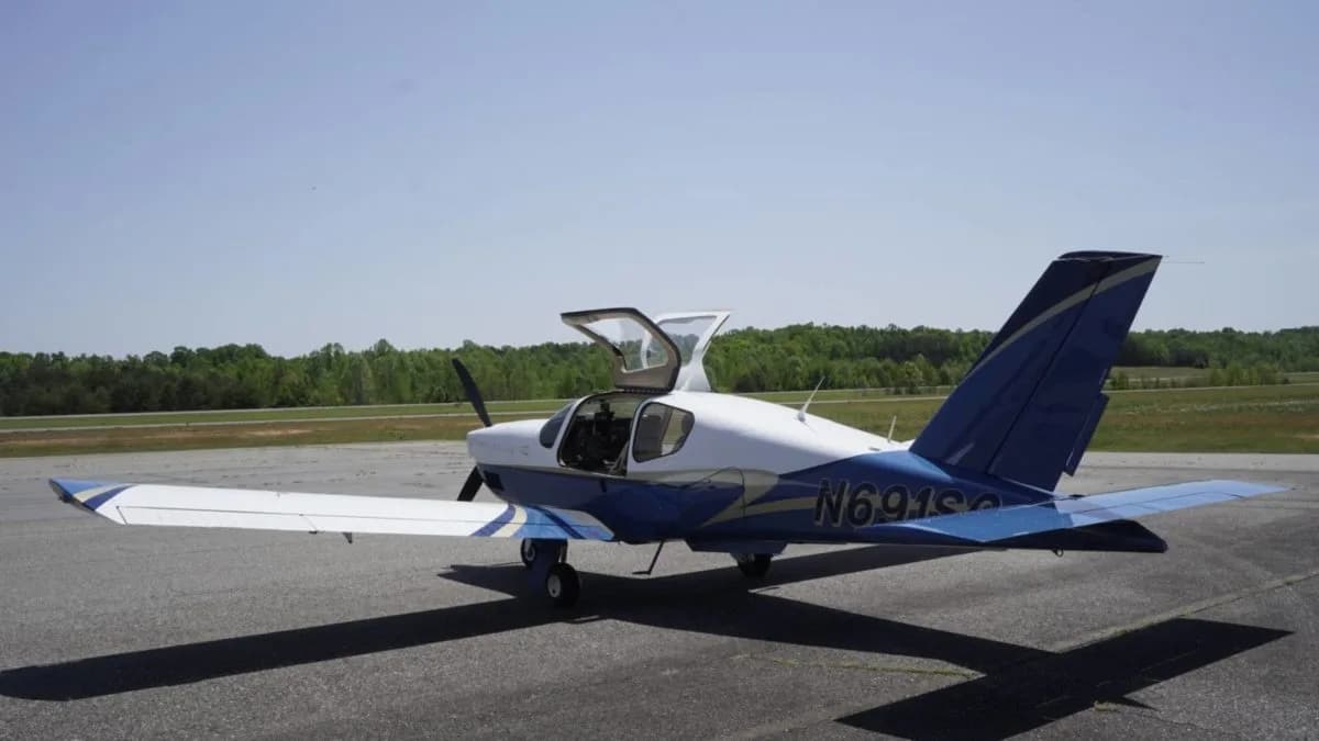 A rear-left view of a white and blue single-engine aircraft with its gull-wing doors open on an airfield.
