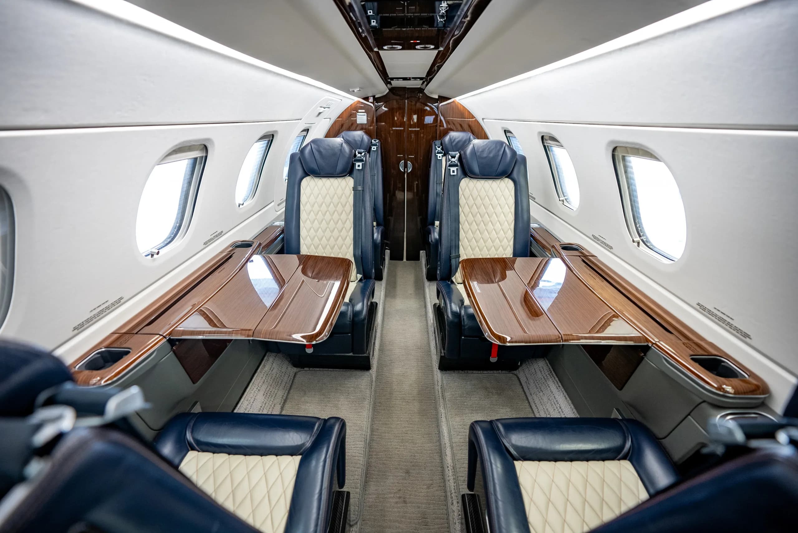 A view down the aisle of the Embraer Phenom 300E cabin, showing opposing dark blue and cream quilted seats with wooden fold-out tables.