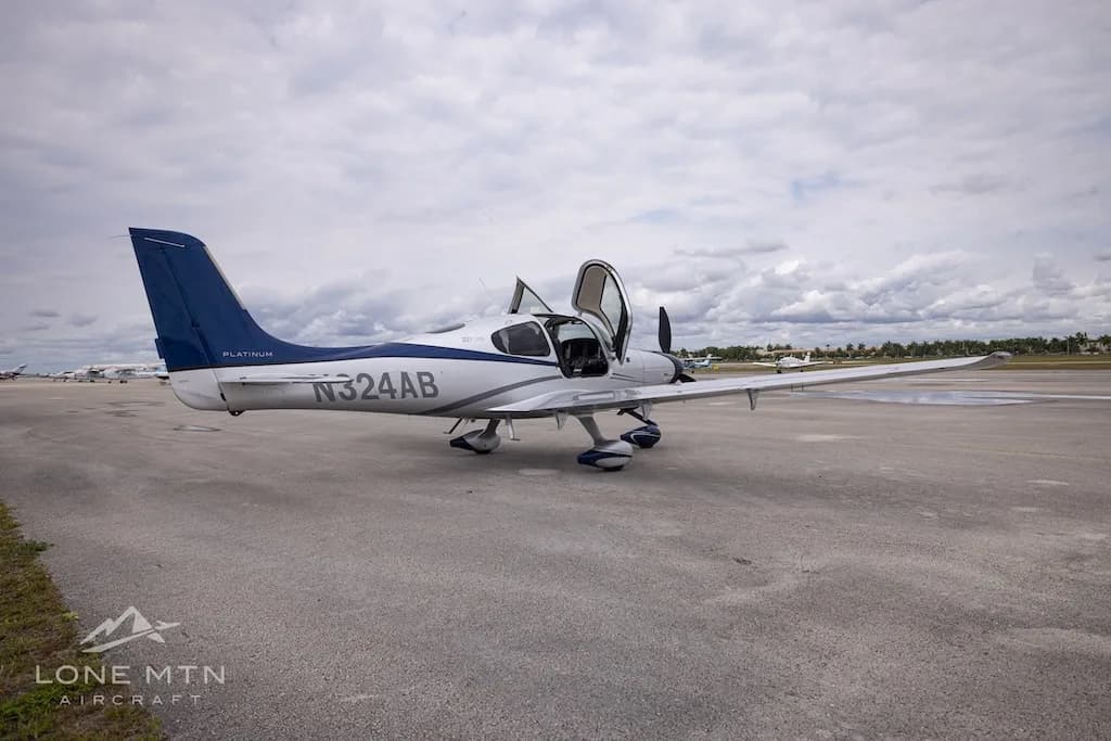 A white and blue Cirrus SR22T aircraft with open doors parked on an airfield, viewed from the rear-right.