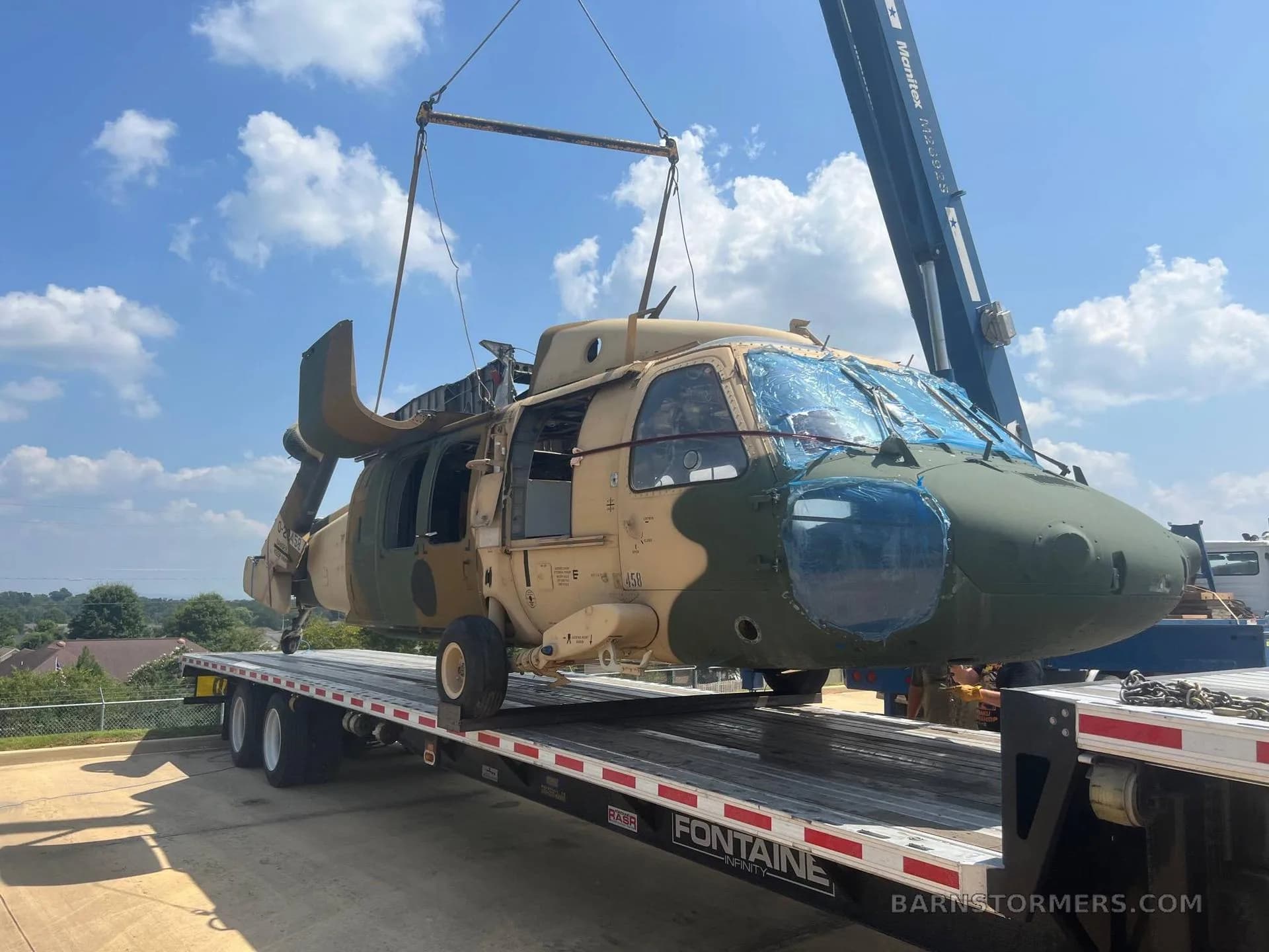 A tan and green camouflage Sikorsky UH-60 Black Hawk helicopter being lifted by a crane onto a flatbed trailer under a blue sky.