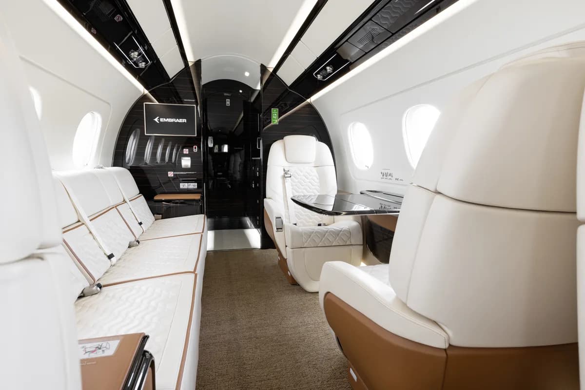 A view inside a private jet cabin showing white quilted seats, a sofa, and a dark wood-paneled rear bulkhead with an Embraer logo.