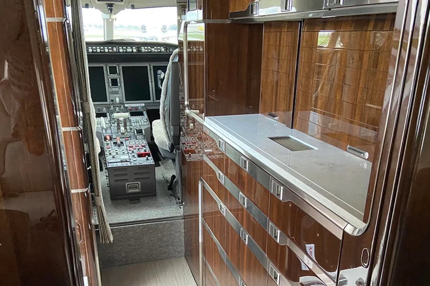 The galley area of a private jet, featuring polished dark wood cabinetry and a countertop, with the cockpit visible in the background.