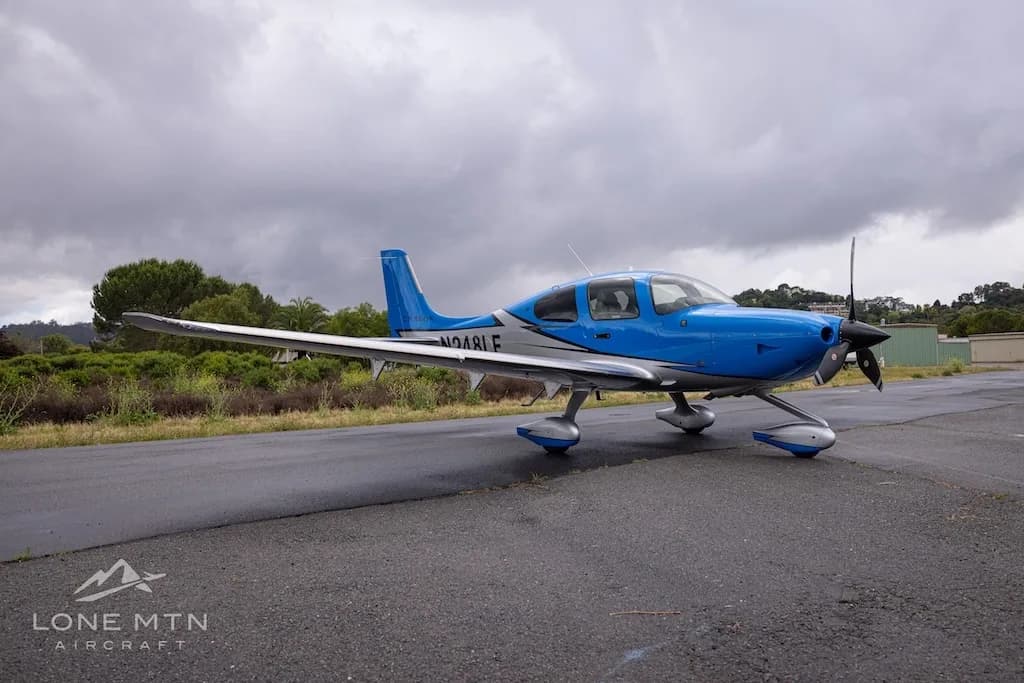 A side view of a blue and silver single-engine propeller aircraft (Cirrus SR22) with registration N248LF parked on a wet tarmac under a cloudy sky.