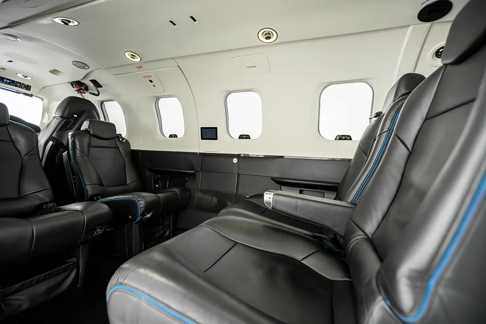 The aircraft cabin interior with black leather seats featuring blue stitching and multiple windows.