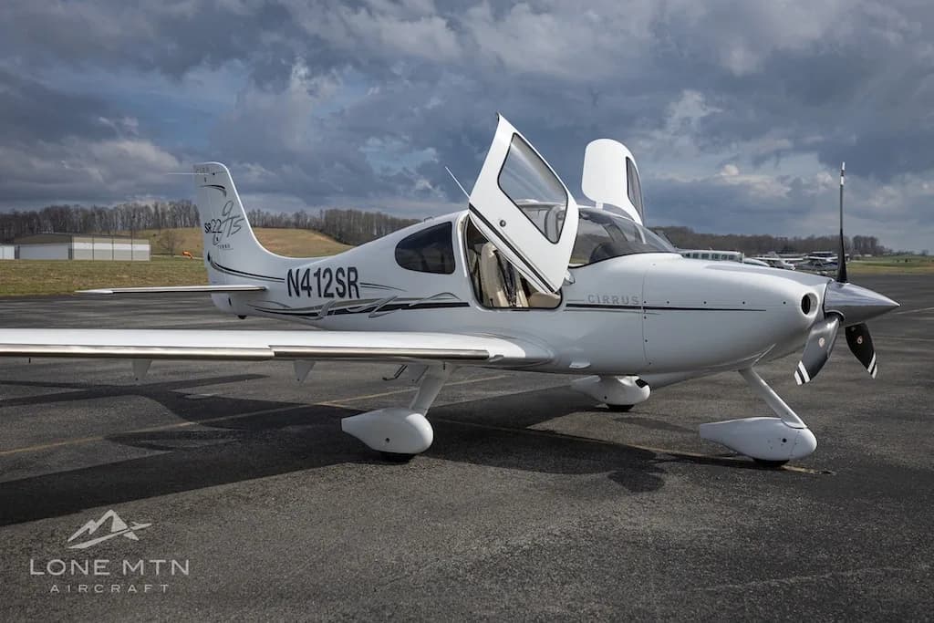 A white Cirrus SR22T aircraft with open doors is parked on an asphalt runway under a dramatic cloudy sky.