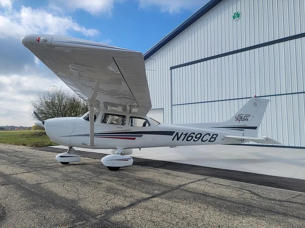 A side view of the white Cessna Skyhawk N169CB with black and red stripes, parked on an asphalt ramp next to a white hangar under a blue sky.