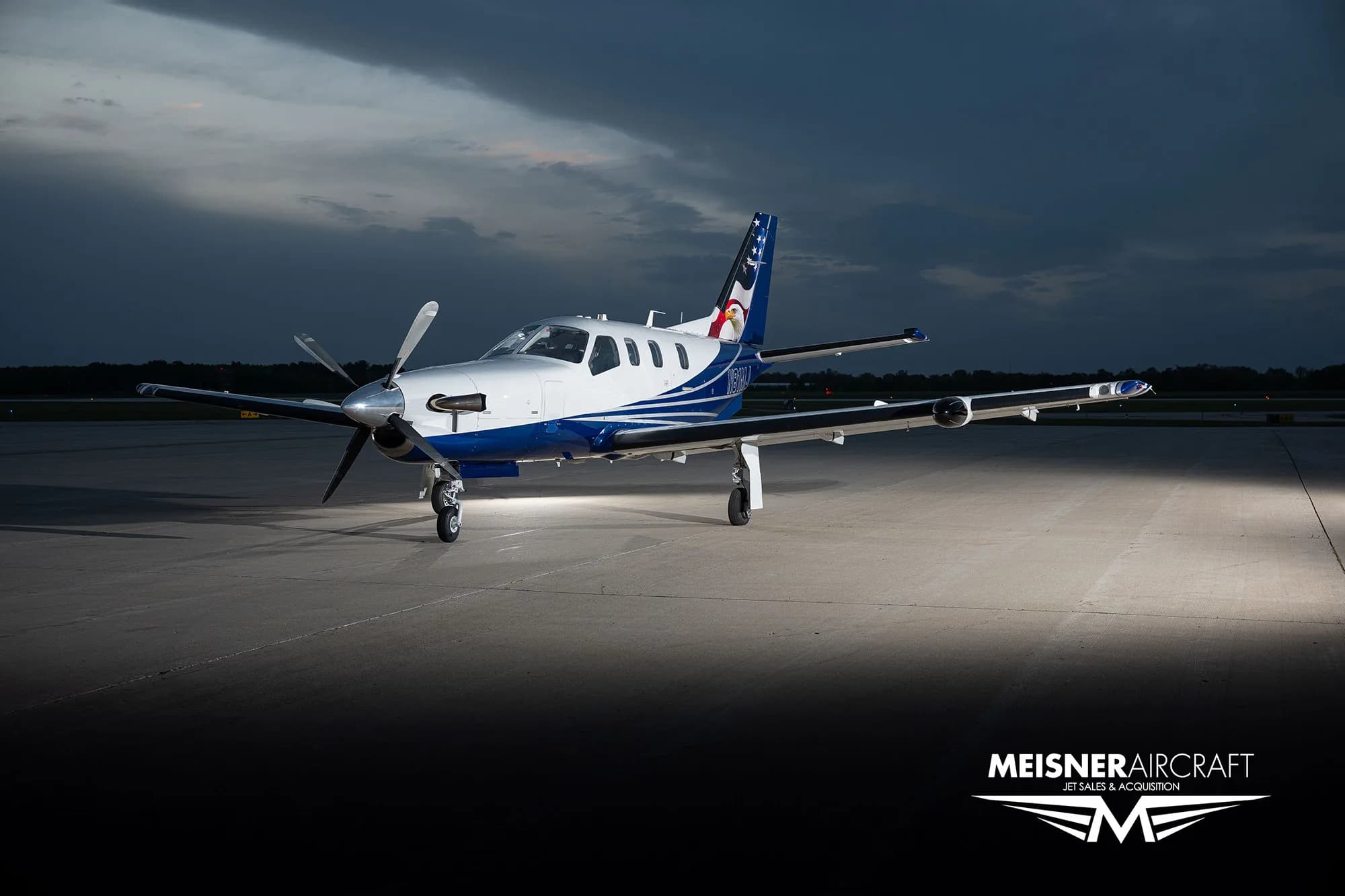 A TBM turboprop aircraft with a blue and white paint scheme and an eagle on the tail, parked on an airfield at dusk, viewed from the front-right.