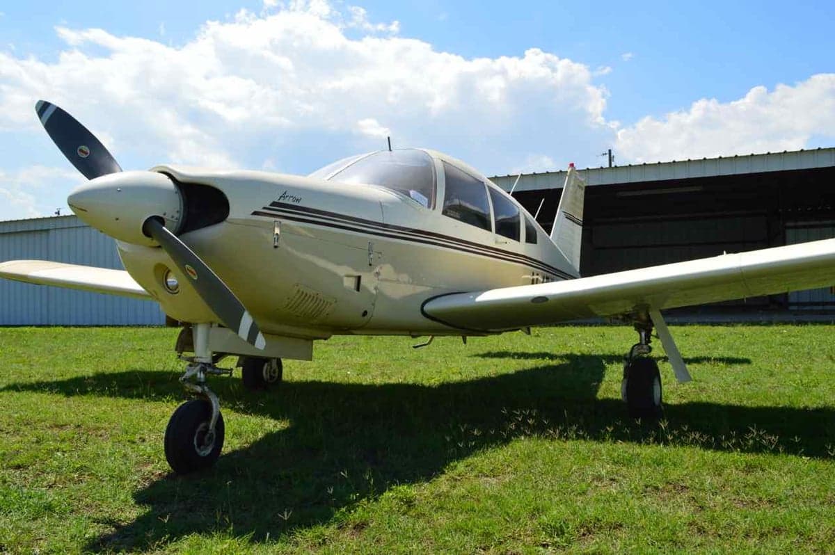 A light-tan Alon Aircoupe sits on a grassy field in front of a hangar.