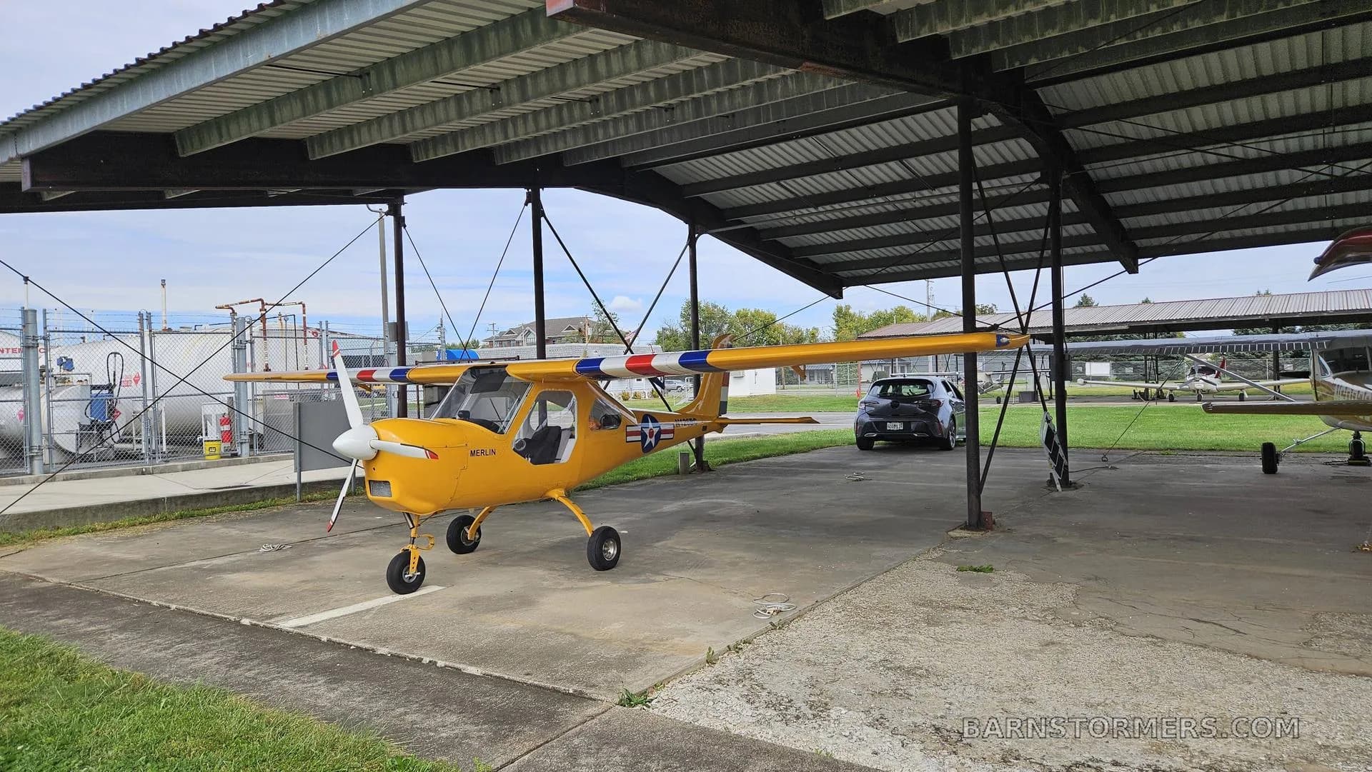 A yellow Merlin ultralight aircraft with a red, white, and blue striped wing is parked under an open-sided hangar, with fuel tanks in the background.