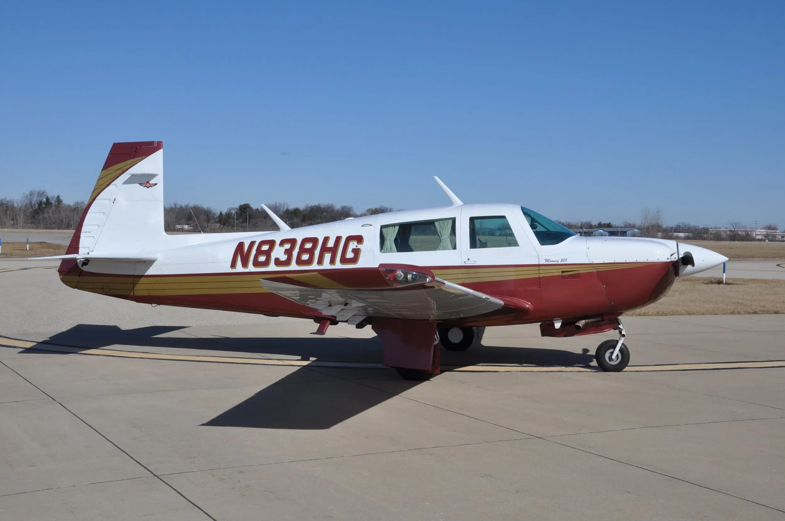 A red and white Mooney 201 aircraft with registration N838HG is parked on an airfield under a clear blue sky.