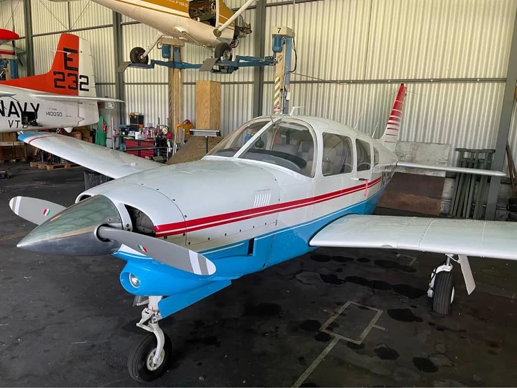 A front-left view of a white and blue Piper PA-28-181 Archer II with red stripes and Italian flag colors on the propeller, inside a hangar.