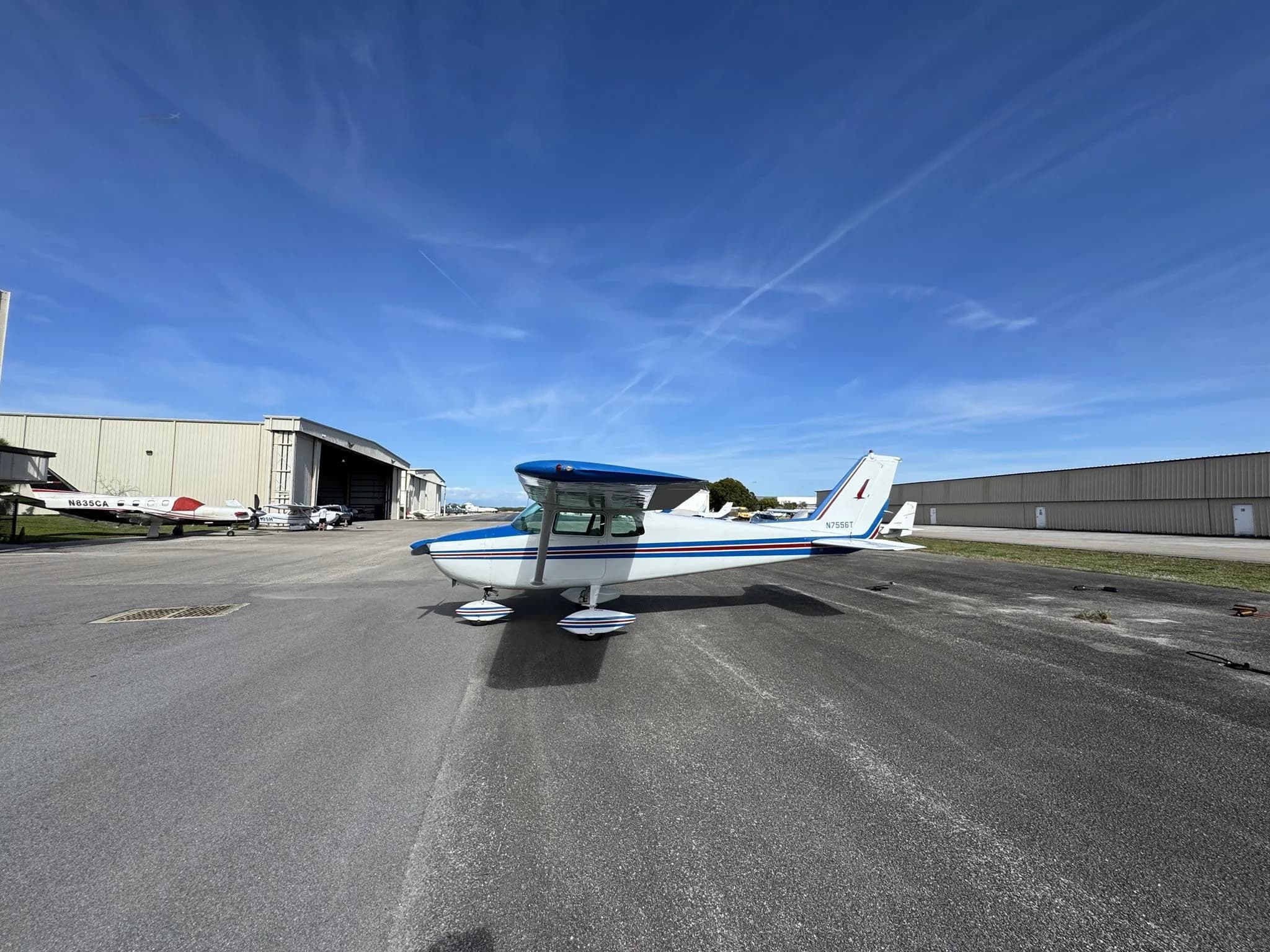 Rear-side view of a white and blue Cessna 172 (N7556T) on an airfield with hangars in the background.