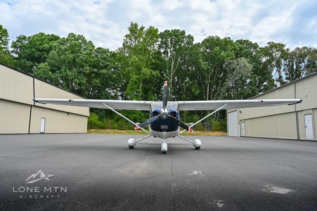 A front view of a blue and white Cessna Skylane N414PK parked between two hangars with trees behind.
