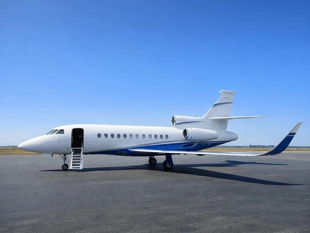 A white and blue Dassault Falcon business jet parked on a tarmac under a clear blue sky, viewed from the side.