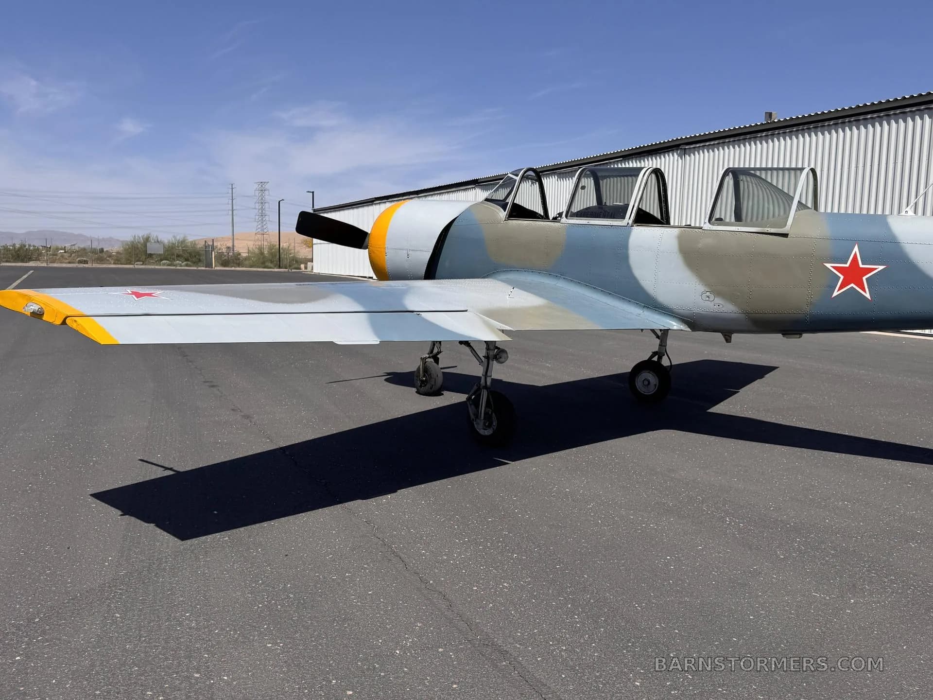 A rear-side view of a blue and grey camouflage Yak-502GC aircraft with a red star on the tail and wing, parked next to a hangar.